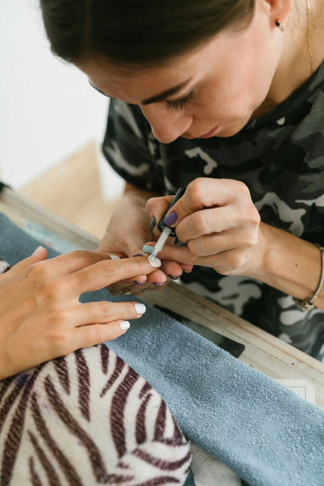 From above of crop concentrated slim ethnic brunette in camouflage T shirt applying white nail polish on nails of unrecognizable woman at home